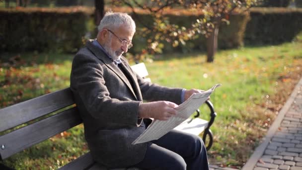 Jóképű nagyapa, gyönyörű szakállal, szürke kabátban ül egy padon a parkban, és újságot olvas. Idősebb ősz hajú férfi szemüvegben — Stock videók