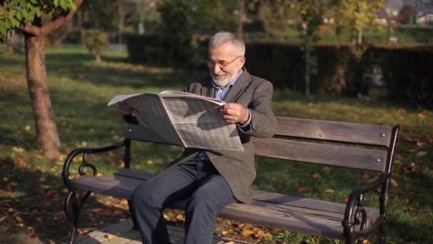 Stilig farfar med vackert skägg i grå jacka sitter på en bänk i parken och läser en tidning. Senior gråhårig man i glasögon — Stockvideo