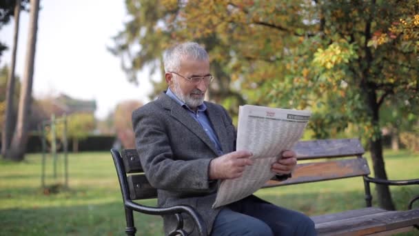 Brodaty starzec w okularach czytający gazetę w jesiennym parku. Przystojny siwowłosy mężczyzna siedzi na ławce wcześnie rano — Wideo stockowe