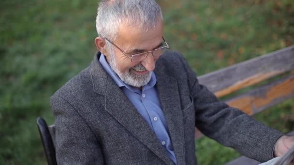 Vousatý starší muž v brýlích čte noviny v podzimním parku. Pohledný šedovlasý muž sedí na lavičce brzy ráno — Stock video