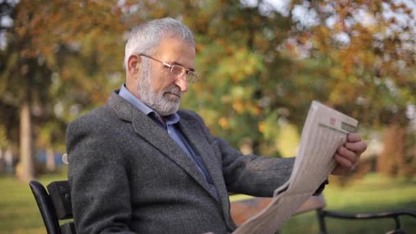 Hombre mayor barbudo en gafas leyendo periódico en el parque de otoño. Hombre guapo de pelo gris sentado en el banco temprano en la mañana — Vídeo de stock