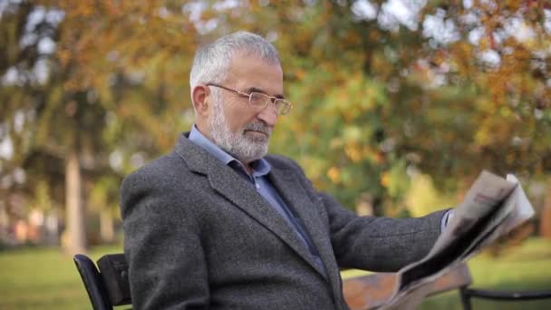 Hombre mayor barbudo en gafas leyendo periódico en el parque de otoño. Hombre guapo de pelo gris sentado en el banco temprano en la mañana — Vídeos de Stock