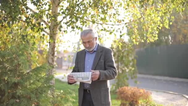 En stilig äldre man går genom parken med tidningen i händerna. Skäggig morfar tidigt på morgonen promenader i parken — Stockvideo