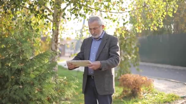 Jóképű végzős férfi sétál a parkban újságpapírral a kezében. Szakállas nagyapa kora reggel sétál a parkban. — Stock videók