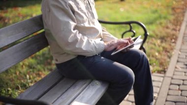 Bankta oturan yakışıklı yaşlı adam internette kaydırma yapmak için tablet kullanıyor. Sonbahar ağacının arkaplanı