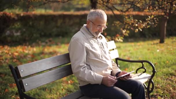 Schöner älterer Mann, der auf der Bank sitzt und mit dem Tablet im Internet blättert. Hintergrund des Herbstbaumes — Stockvideo