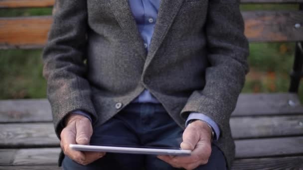 Handsome gray-haired elderly man with beautiful beard sitting on the bench and use tablet for scrolling in internet. Background of autumn tree — Stock Video