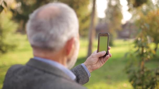Изображение пожилого человека, использующего телефон на улице. Зеленый экран. Задний вид человека, держащего телефон на неровной длине. Дедушка снимал видео на смартфоне — стоковое видео