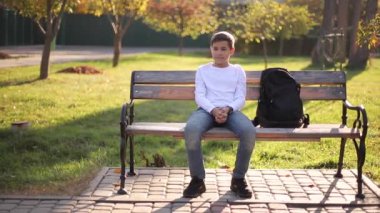 Bankta oturan beyaz kazaklı genç çocuk. Sonbahar parkında sırt çantalı yakışıklı okul çocuğu.