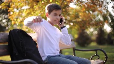 Beyaz tişörtlü, siyah sırt çantalı, parkta bankta oturan ve telefonla konuşan genç bir çocuk.