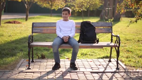 Tonårspojken i vit tröja sitter på bänken. Snygg skolpojke med ryggsäck i höstparken — Stockvideo