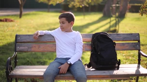 Tienerjongen in witte trui zittend op de bank. Knappe schooljongen met rugzak in het herfstpark — Stockvideo