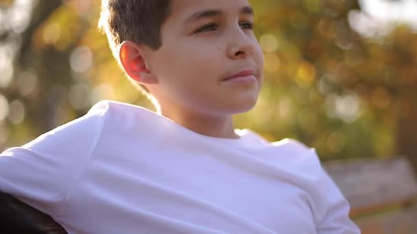 Gros plan du garçon adolescent en pull blanc assis sur le banc. Beau garçon d'école avec sac à dos dans le parc d'automne — Video