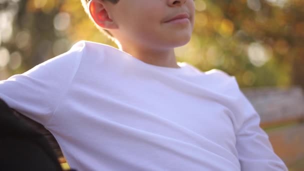 Gros plan du garçon adolescent en pull blanc assis sur le banc. Beau garçon d'école avec sac à dos dans le parc d'automne — Video