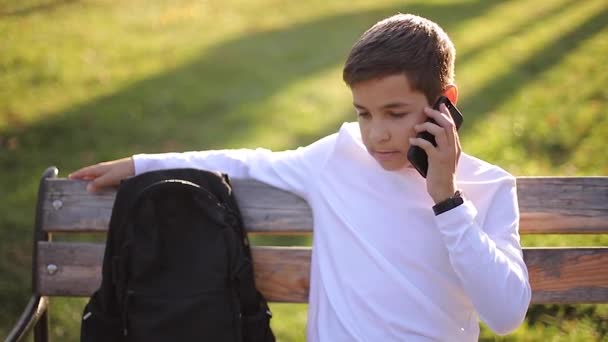 Мальчик в белой толстовке с черным рюкзаком сидит на скамейке в парке и разговаривает с кем-то по телефону — стоковое видео