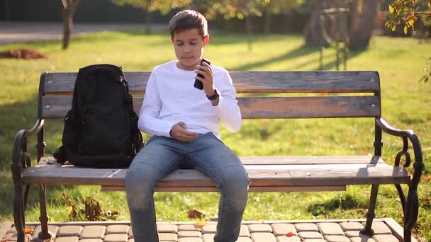 Niño en sudadera blanca con mochila negra sentado en el banco en el parque y hablar con alguien por teléfono — Vídeo de stock