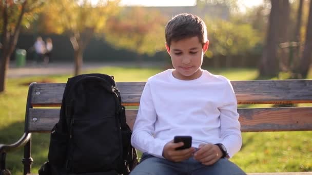 Хлопчик-підліток грає в онлайн ігри на своєму смартфоні в парку після уроків у школі — стокове відео