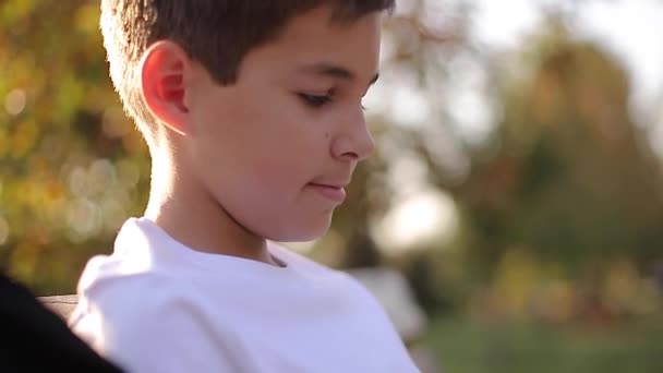 Gros plan du garçon adolescent en pull blanc assis sur le banc. Beau garçon d'école avec sac à dos dans le parc d'automne — Video