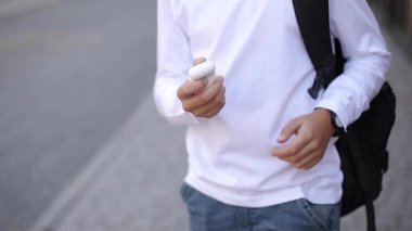 Genç çocuk kablosuz kulaklığı çıkarıp kulaklıklara takıyor. Sırt çantalı beyaz gömlekli genç bir çocuk. Çocuk müziği dinle.