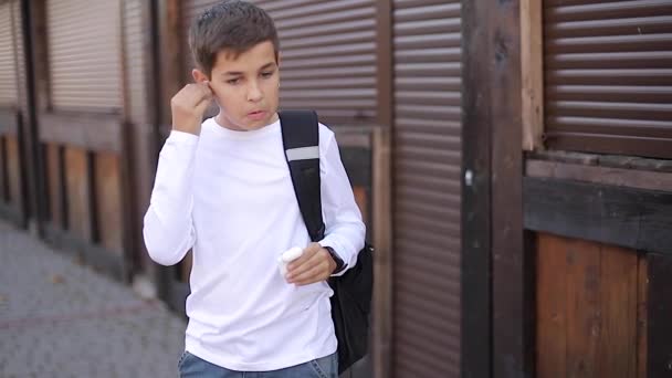 Teenage boy take out a wireless headphones and put then into the ears. Young boy in white shirt with backpack. Boy listen music — Stock Video