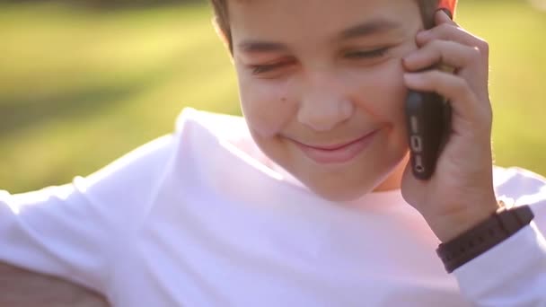 Young boy in white sweatshirt with black backpack sitting on the bench in the park and speak with somebody by the phone — Stock Video