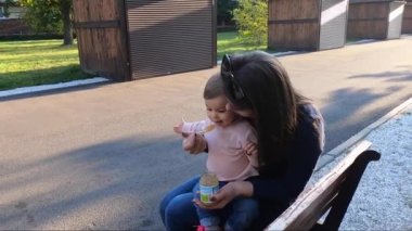 Küçük kız parkta yemek yiyor, anne de bebek meyvelerini veriyor. Anne ve kızı bankta oturuyor.