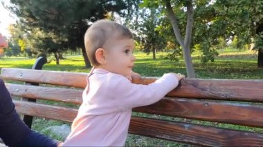 Küçük kız parkta yemek yiyor, anne de bebek meyvelerini veriyor. Anne ve kızı bankta oturuyor.