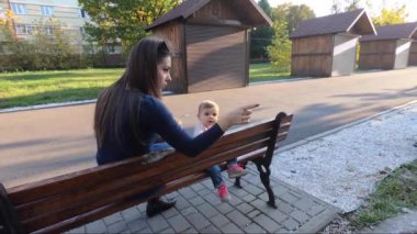 Küçük kız parkta yemek yiyor, anne de bebek meyvelerini veriyor. Anne ve kızı bankta oturuyor.