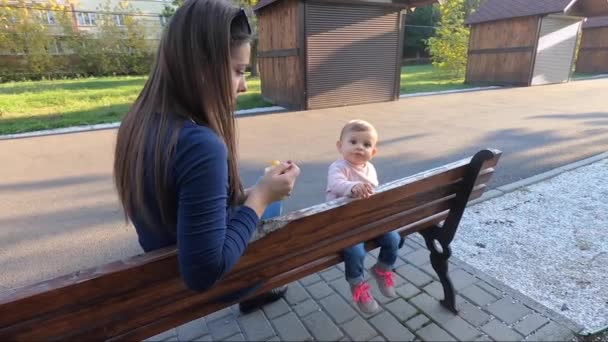 En liten flicka äter i parken, mamma ger till babyfrukter. Mamma och dotter sitter på bänken — Stockvideo