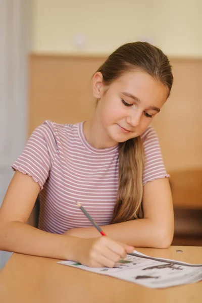 Happy schoolgirl sitting at desk and writing in exercise book — Stock Photo, Image