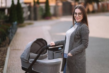 Aile, çocuk ve ebeveyn konsepti-parkta puset ile mutlu anne. Güneş gözlüğü ve şık gri ceket ile anne