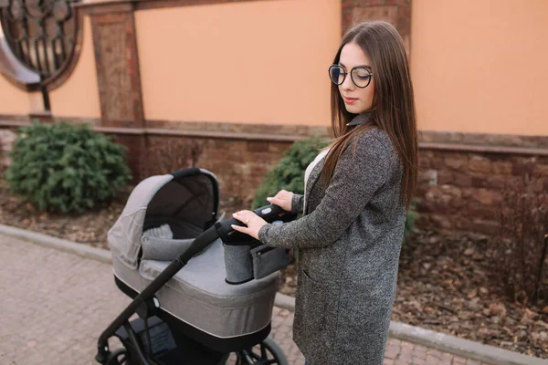 Madre joven caminando con su bebé recién nacido en cochecito. Un cochecito gris. Hermosa mamá en gafas de sol — Foto de Stock