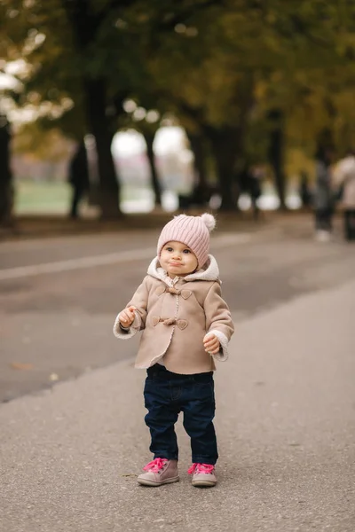 11 개월된 아기가 공원을 걷고 있습니다. 귀여운 갈색 코트에 핑크 모자와 데님 청바지를 입은 행복 한 아기 — 스톡 사진