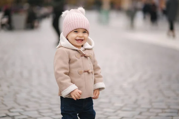 Klein glimlachend meisje gekleed in bruine jas denim jeand en roze hoed staan in het centrum van de stad. Schattig meisje. — Stockfoto