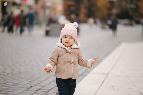 Klein glimlachend meisje gekleed in bruine jas denim jeand en roze hoed staan in het centrum van de stad. Schattig meisje. — Stockfoto