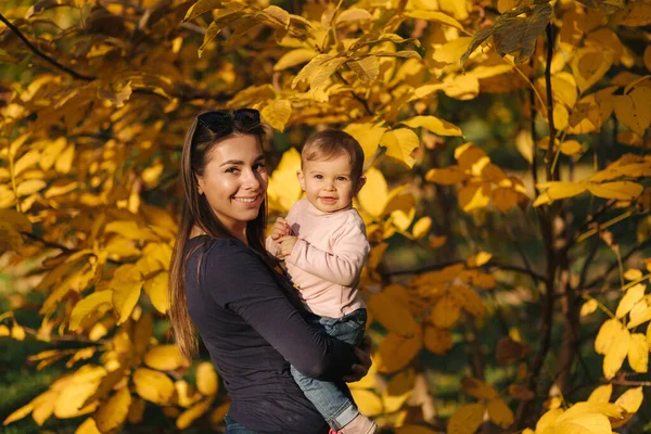 Ritratto di bella famiglia nel parco. Mamma e figlia insieme in autunno. Bellissimo bambino di dieci mesi sulle mani delle madri. Sfondo dell'albero giallo — Foto Stock