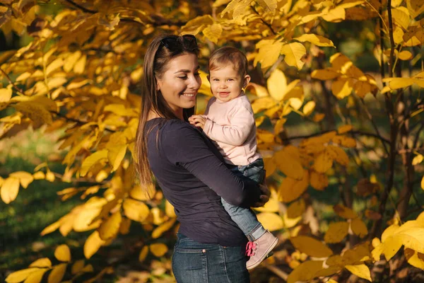 Porträtt av vacker familj i parken. Mamma och dotter tillsammans på hösten. Vacker tio månaders bebis på mammas händer. Bakgrund av gult träd — Stockfoto