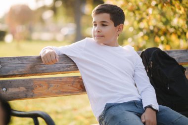 Bankta oturan beyaz kazaklı genç çocuk. Sonbahar parkında sırt çantalı yakışıklı okul çocuğu.
