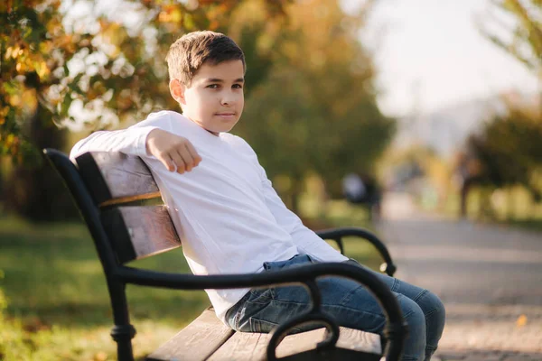 Nastolatek w białym swetrze siedzi na ławce. Przystojny chłopak ze szkolnym plecakiem w jesiennym parku — Zdjęcie stockowe