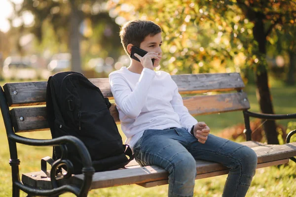 Niño en sudadera blanca con mochila negra sentado en el banco en el parque y hablar con alguien por teléfono —  Fotos de Stock