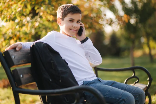 Kleiner Junge in weißem Sweatshirt mit schwarzem Rucksack sitzt auf der Parkbank und telefoniert mit jemandem — Stockfoto