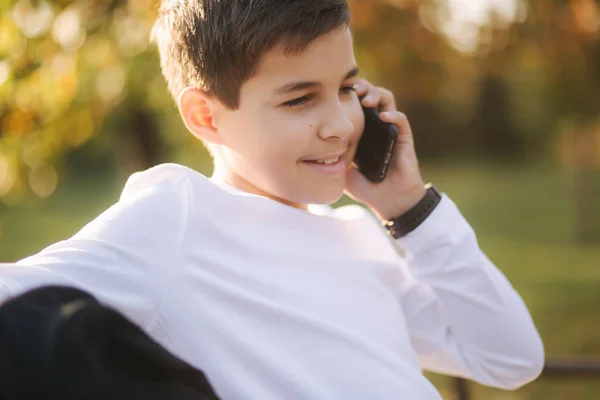 Niño en sudadera blanca con mochila negra sentado en el banco en el parque y hablar con alguien por teléfono — Foto de Stock