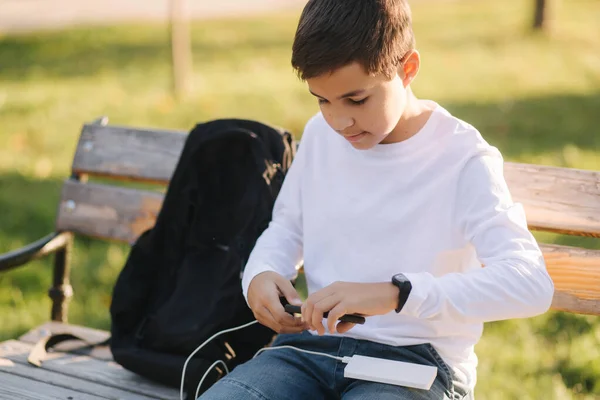 白いシャツを着た10代の少年は、スマートフォンを充電するためにパワーバンクを使用しています。スマートフォンの低電池 — ストック写真