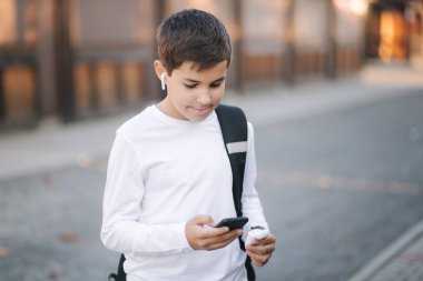 Beyaz tişörtlü, siyah sırt çantalı, telefon, kablosuz kulaklık ve sosyal ağda yazılı mesaj kullanan genç bir çocuk.