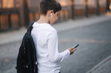 Beyaz tişörtlü, siyah sırt çantalı, telefon, kablosuz kulaklık ve sosyal ağda yazılı mesaj kullanan genç bir çocuk.