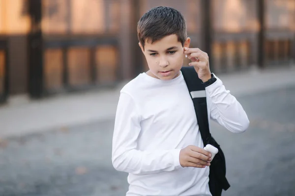 Tienerjongen pakt een draadloze koptelefoon en stopt die in de oren. Jonge jongen in wit shirt met rugzak. Jongen luister muziek — Stockfoto