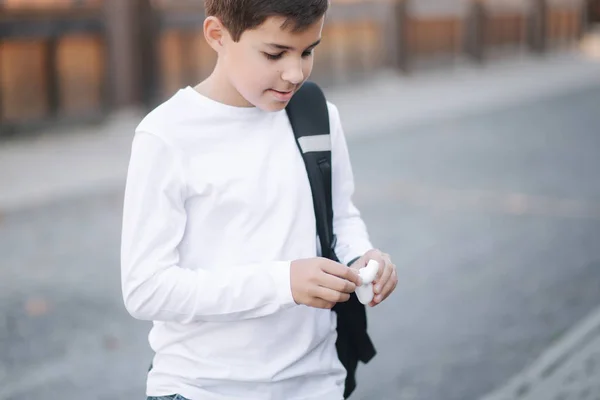 Tonårspojken tar fram en trådlös hörlurar och sticker in den i öronen. Ung pojke i vit skjorta med ryggsäck. Pojke lyssna musik — Stockfoto