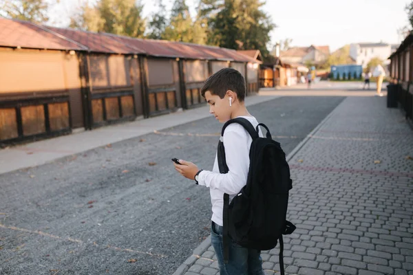 白のスウェットシャツと黒のバックパック付きの10代の男の子は、ソーシャルネットワークで携帯電話やワイヤレスヘッドフォンや印刷メッセージを使用しています — ストック写真