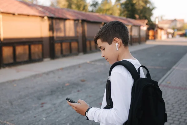 Menino adolescente em camisola branca e com backpuck preto usar telefone e fones de ouvido sem fio e imprimir mensagem em rede social — Fotografia de Stock