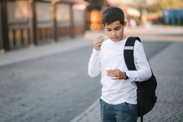 Nastolatek wyciąga słuchawki bezprzewodowe i wkłada je do uszu. Młody chłopak w białej koszuli z plecakiem. Chłopiec słuchać muzyki — Zdjęcie stockowe